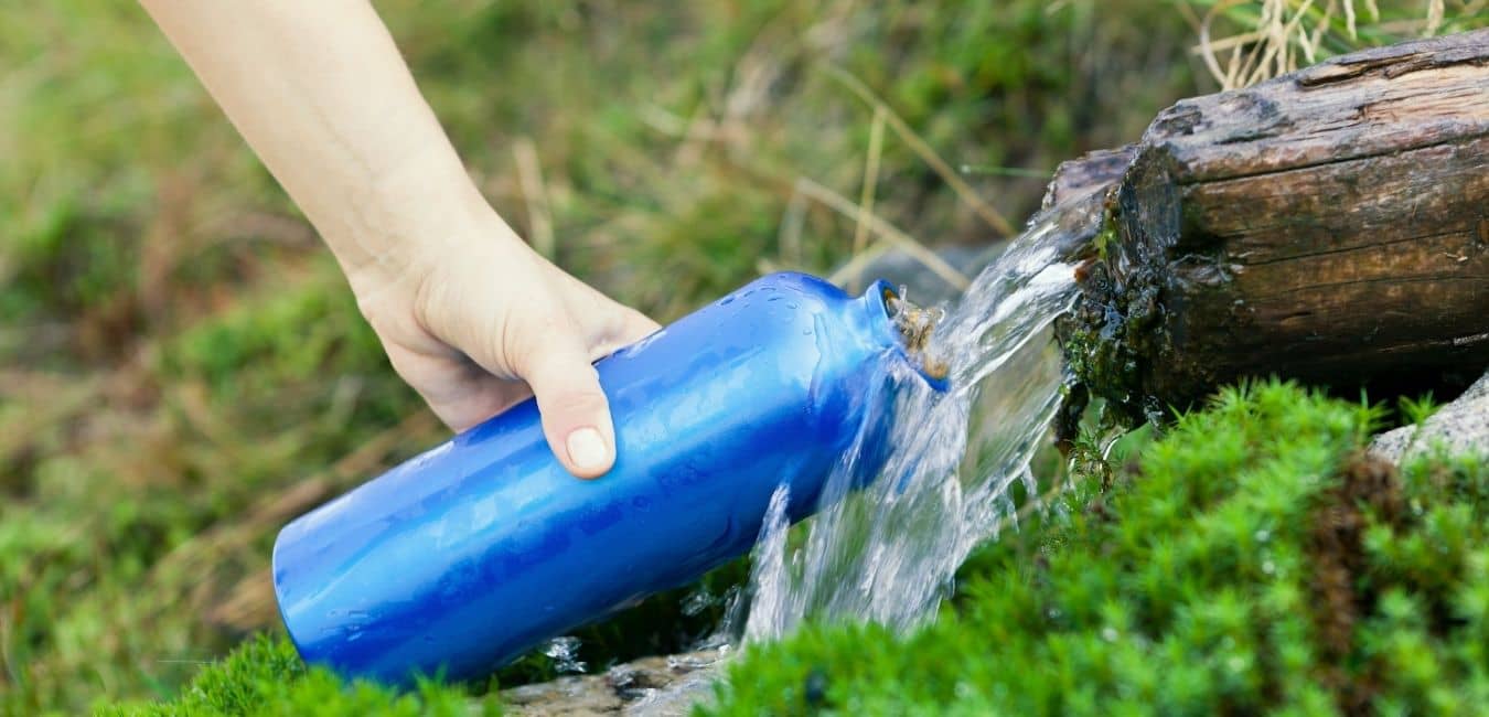 Best hydration bladder and hiking tips. Woman holding blue water bottle under flowing water during a hike.