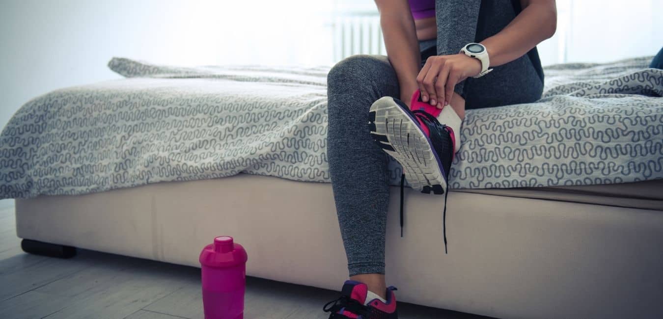 How to get fit. Woman sitting on edge of bed in workout clothes putting on pink and gray gym shoes with a pink water bottle sitting on floor.