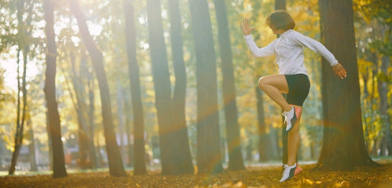 The Best & Worst Times to Start Working out. Woman doing high knee raises in forest with sun peaking through trees.