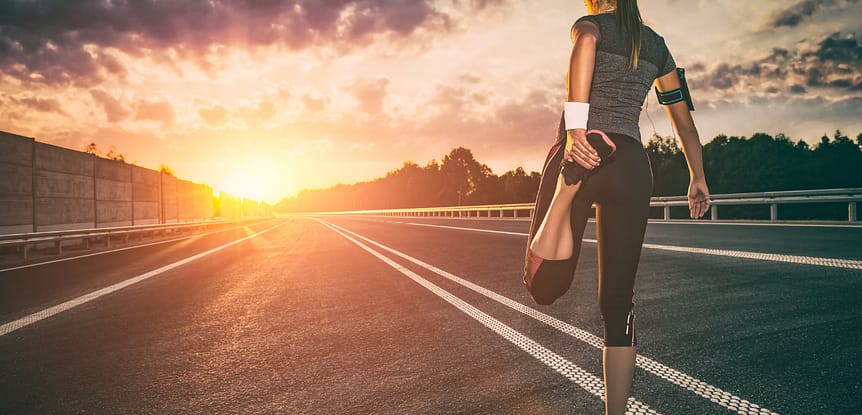 A Workout Plan You Can Stick to. Woman standing on a road stretching her quads getting ready for a run looking at a sunset.