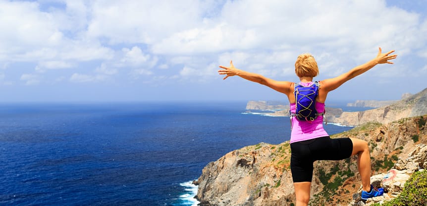 What Goals Should I Set and how do I achieve them? Woman with pink shirt and purple daypack standing on mountain overlooking ocean with arms stretched to sky.