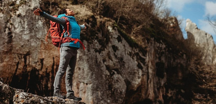 Freedom Is Fit, Strong, Confident, Adventurous and Pain-Free Joints! Woman with arms outstretched on a mountain top wearing a blue coat and orange backpack.