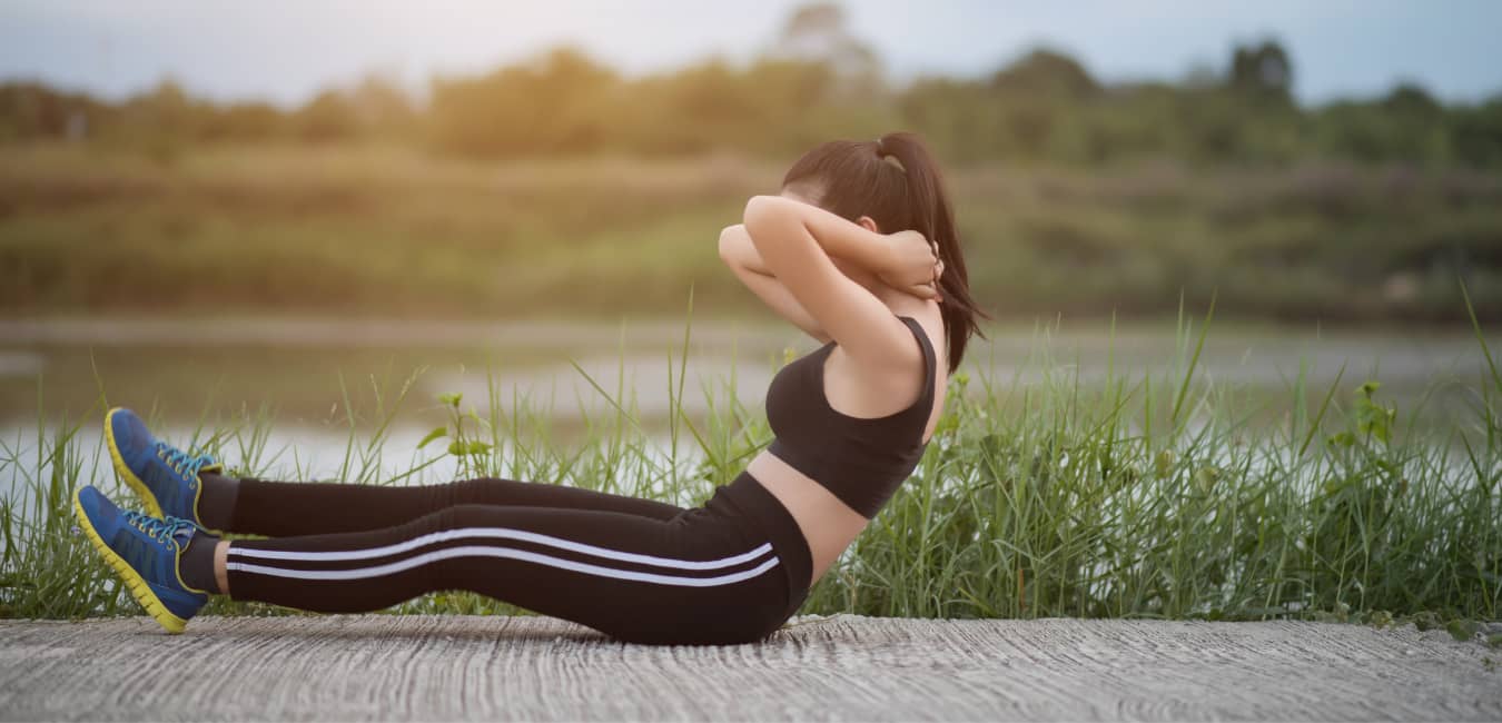 Outdoor workout, no equipment required. Woman sitting outdoors doing sit ups. Grassy background, paved surface.
