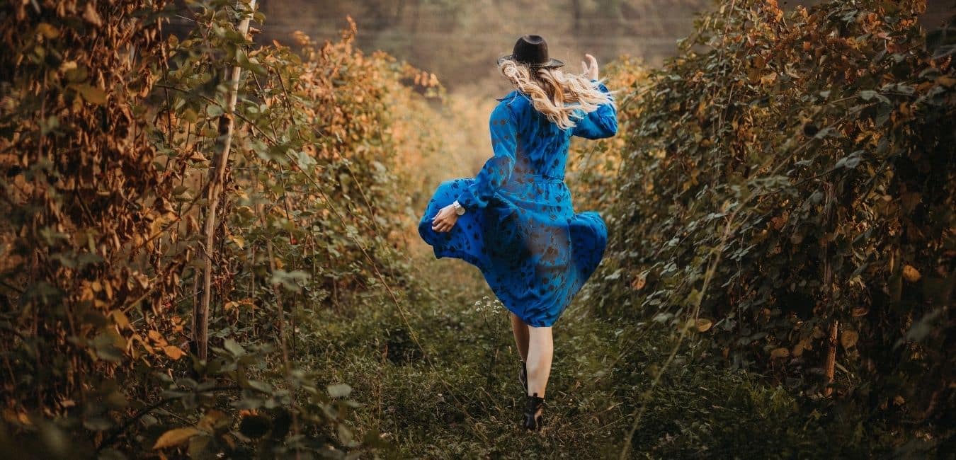 She was having so much fun, her family was worried. Woman in a blue dress and black hat skipping through a field.