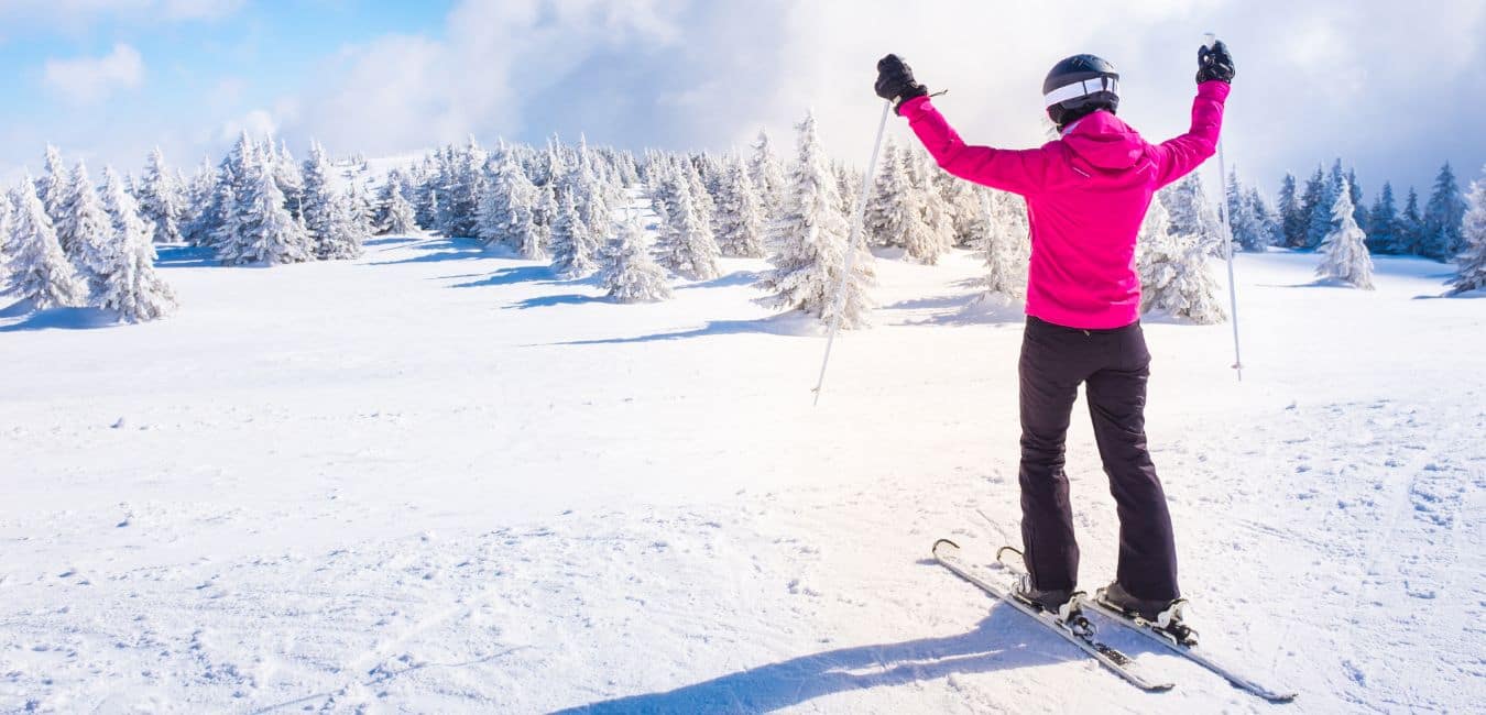 December can be a Month of Fitness Fun! woman in pink coat and skis with arms raised.
